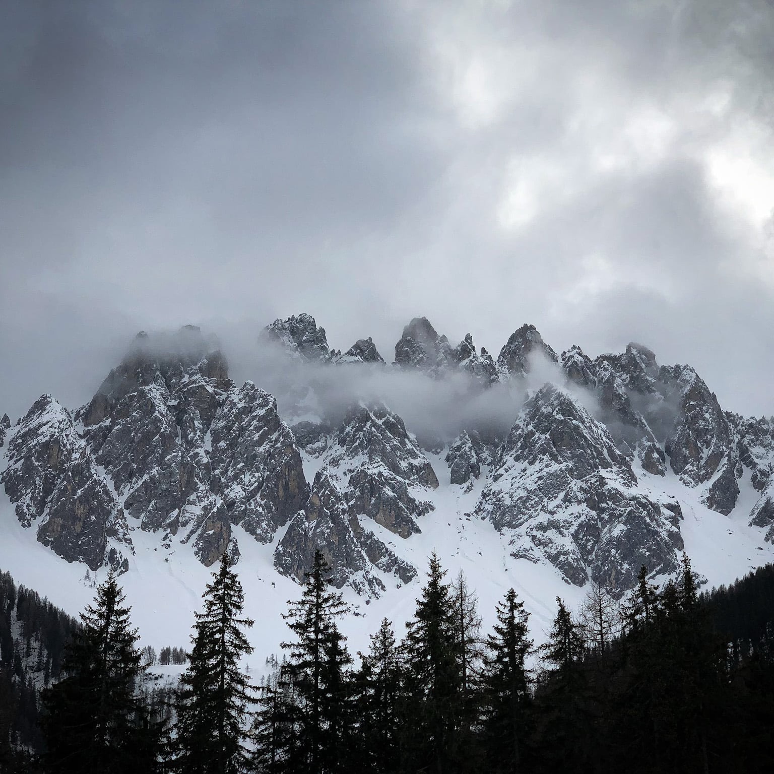 Snowy peaks among the clouds, above a dark forest.