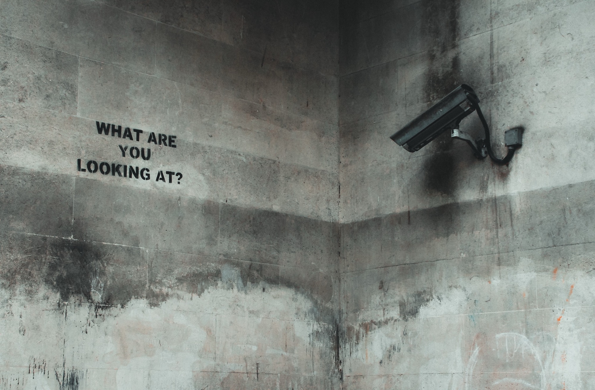 A security camera and the words “What are you looking at?” on a concrete wall.